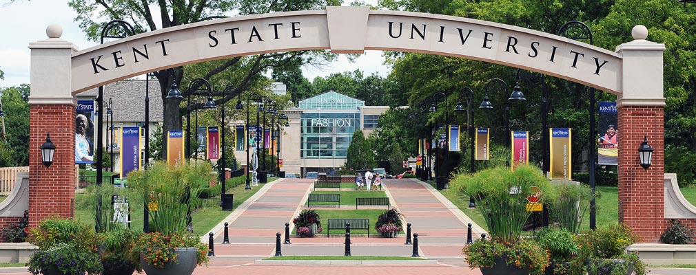 Photograph of an archway with the words “Kent State University” at the start of the esplanade, with two wide walkways; benches and lights; and lawns, bushes, and mature trees. A multistory university building ends the vista down the esplanade.