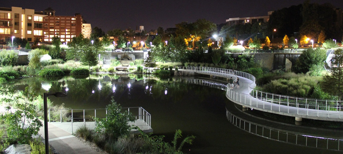 The photograph shows a picture of the park in the evening; a walking trail follows a water feature and greenspaces are lit during the evening.