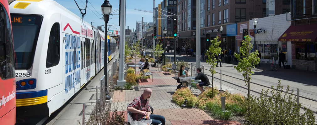 View of a downtown area with a light rail train stopped along a wide median with benches and tables and chairs occupied by people.