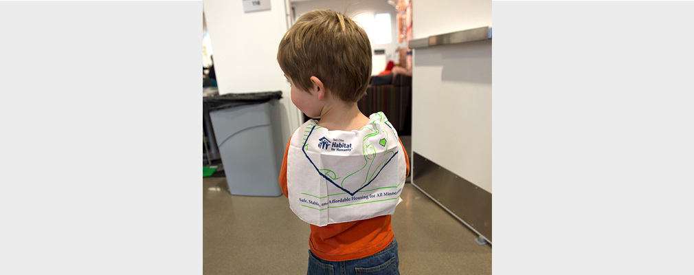 A little boy is seen from behind with a Habitat for Humanity cloth poster wrapped around his shoulders.