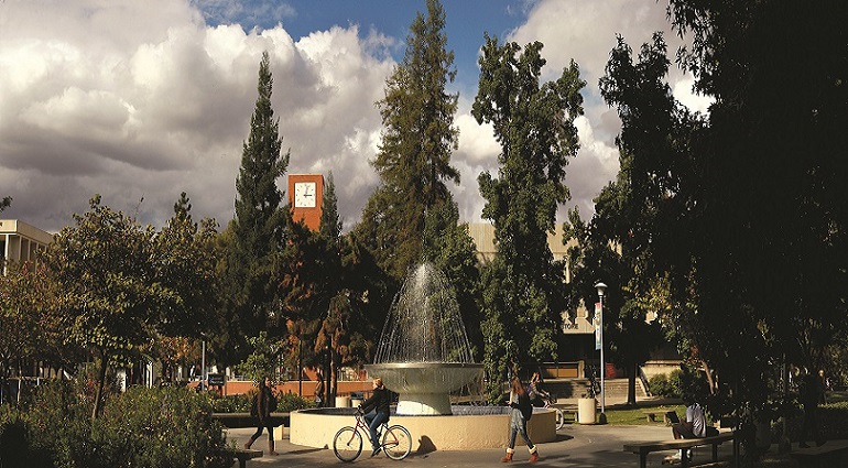 Photograph of students walking, bicycling, and sitting near a fountain surrounded by vegetation as they travel across the campus of California State University at Fresno. A clock tower and campus buildings are in the background behind tall trees.