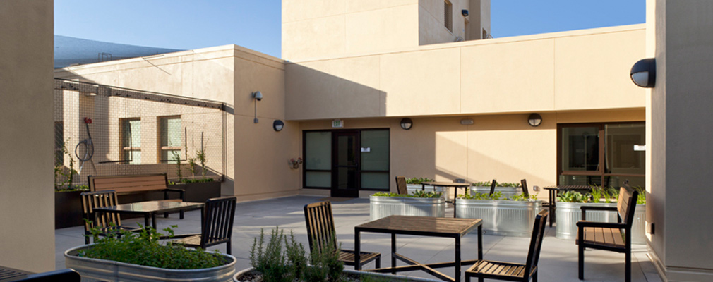 Photograph of tables, benches, and planters in a courtyard with the walls of the ninth floor rooms in the background.