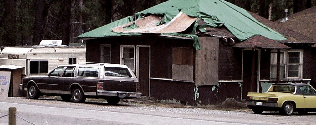 Photograph of a dilapidated, one-story wooden building. Portions of its walls are covered with plywood panels. Sections of the pyramidal roof are missing, covered by torn tarps.