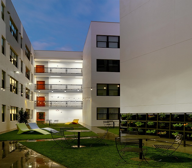 An evening photograph of the courtyard framed by the four residential stories of the apartment building. The courtyard is grassed, with concrete sidewalks. Three lounge chairs and four bistro tables with chairs stand in the grassed areas. Strings of lights are hung overhead.