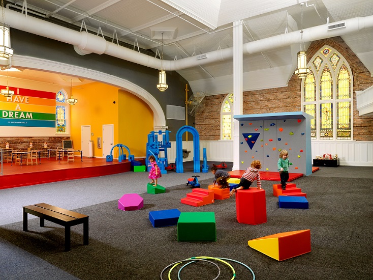 A photograph of the play space inside the church. Four preschoolers play on brightly colored geometric forms in the middle ground. Other play equipment stands behind the children. A bench and several hoola-hoops are on the floor in the foreground. To the rear, the play area is framed by a stage and an exterior wall of the church, with exposed brick and two stained glass windows.