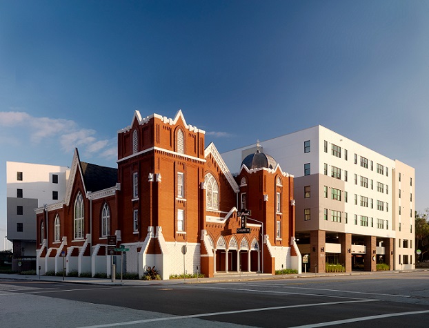 Photograph taken at street level of the Metro 510 development on the opposite corner of an intersection. The photograph shows the front and side facades of the St. James A.M.E. Church, a brick neoGothic building at the corner. To the side and rear of the church is the apartment building, with four residential floors above a two-story parking garage; the facades of the apartment building are very plain, with no ornamentation, serving as a backdrop to the historic church.