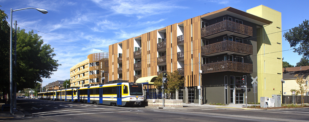 Photograph showing a light rail train stopped in front of four-story apartment buildings.