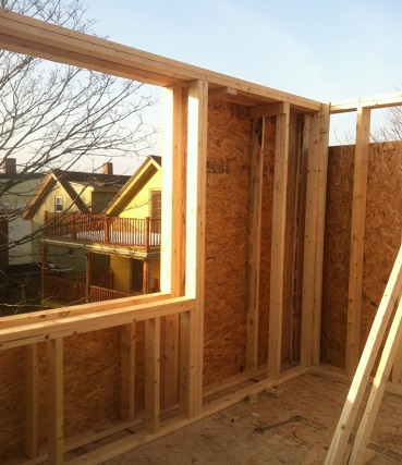 Photograph taken from an upper floor of one of the townhouses during construction. The photo shows the wood framing of the double stud walls and an open sky above. There is a gap in the framing for a window on the left side of the photograph with a view of two houses in the background. 