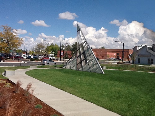 Photograph of a metal and glass sundial art piece approximately 10 feet tall standing in a lawn and encircled by a sidewalk. A large brick building and a smaller structure with siding, as well as a street with parked cars, are in the background.