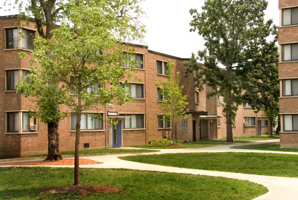 Ground-level photograph of a three-story brick building in Parkway Gardens. In front of the building are paved walkways, young and mature trees, and a lawn. A corner of a second building is at the right-hand edge of the photograph.
