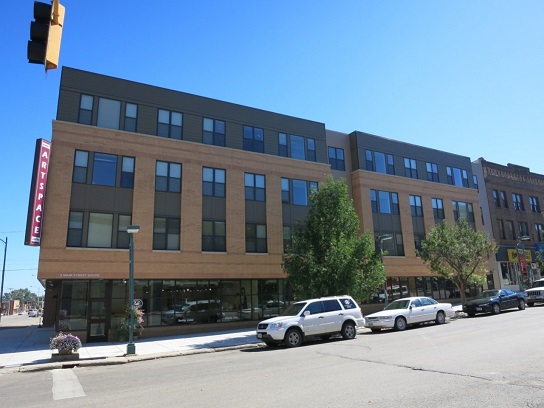 Photograph from across the street of Minot Artspace Lofts building, showing the four-story brick building on a corner lot. A vertical sign with the words “Minot ARTSPACE Lofts” is projected from the front corner of the building. A wide sidewalk, street trees, and cars parked along the curb are visible in the foreground.