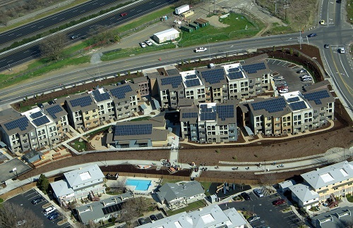 Low-angle aerial photograph of 3-story buildings and a 1-story community building comprising the New Harmony development. Arrays of solar panels are visible on all the roofs. Also in the picture are abutting streets and apartments. 