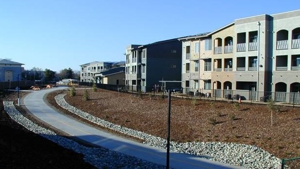 Picture taken just off site from ground level showing the facades of several of the 3-story apartments and the community building in New Harmony. In the foreground is the bicycle path constructed as part of the development.