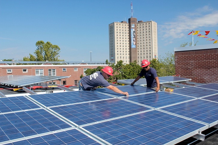 Photograph of two workers affixing a solar panel to an array of panels on the flat roof of a midrise building. The tops of a midrise and a high rise building are in the background.