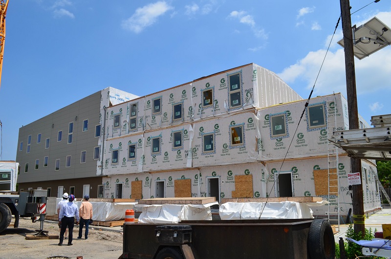 Photograph at ground level of the front of the eight-unit building under construction. Exterior siding has been applied to four of the townhomes. The other townhomes do not have exterior siding, and the modular construction is evident; the end townhome has only two of its three modules in place. Construction materials and equipment and three workers are in the foreground of the picture.