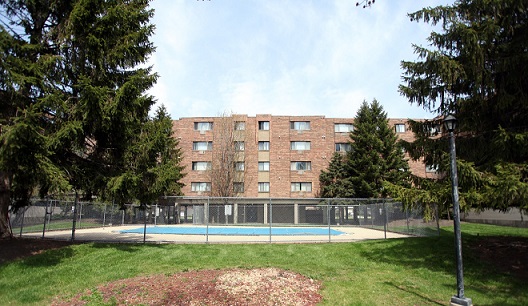 Image displaying the rear side of a multistory, brick residential building (2000 Illinois), as well as a covered swimming pool, enclosed by a fence and surrounded by grass and large trees.