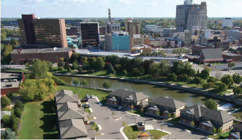 Photograph taken of downtown Flint, Michigan. Six low-lying buildings in the foreground lie adjacent to a river that runs through the center of the photograph. A variety of low- and mid-rise buildings can be seen in the background of the photograph.