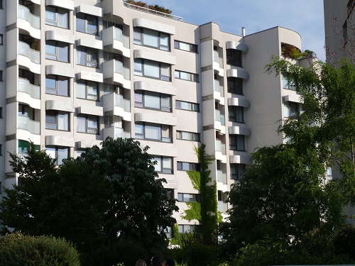 Photograph taken at street level showing a façade of one of the buildings of the Wohnpark Neue Donau housing community. The housing units are shown to have balconies and large windows. Trees in the foreground beautifully frame the view of this seven-storied building.