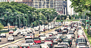 Cars travel along a congested/busy multi-lane divided road, surrounded by street trees and multi-story buildings.