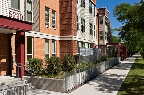 Exterior image of a multistory residential building, with a mixed brick and siding façade. A sidewalk and trees are visible in front of the building.