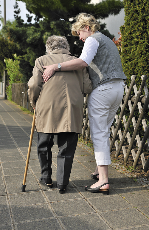An older woman walking down a sidewalk, aided by the use of a cane and a walking companion.