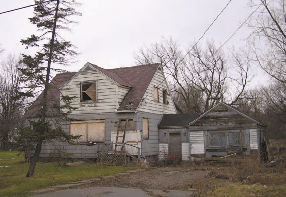 A dilapidated, tax-foreclosed single-family home on a weedy and junk-strewn lot with broken, boarded-over windows and remnants of a concrete driveway.