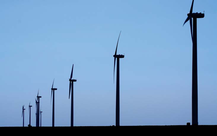 A row of wind generators spread across a field, backlighted by the sky.