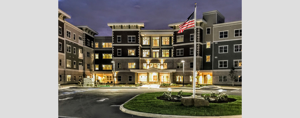Photograph at night of the main entrance to a 4-story multifamily building, with an American flag on a pole in the foreground.