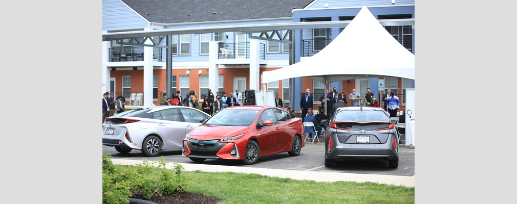 Photograph of a crowd gathered beneath a temporary pavilion and three hybrid vehicles, with a two-story residential building in the background. 