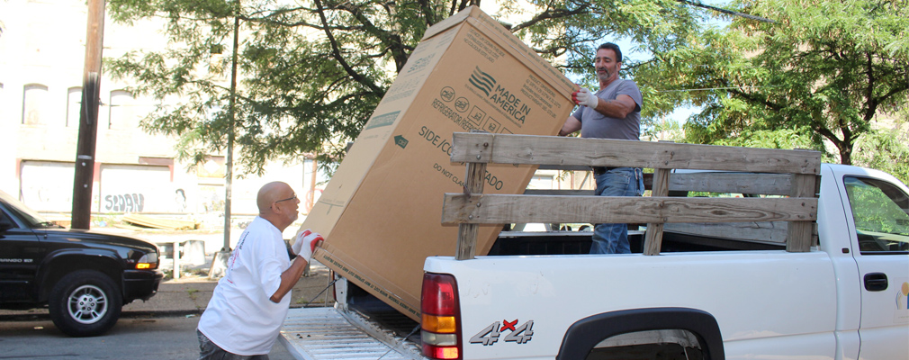 Photograph of two men unloading a boxed refrigerator from a pickup truck.