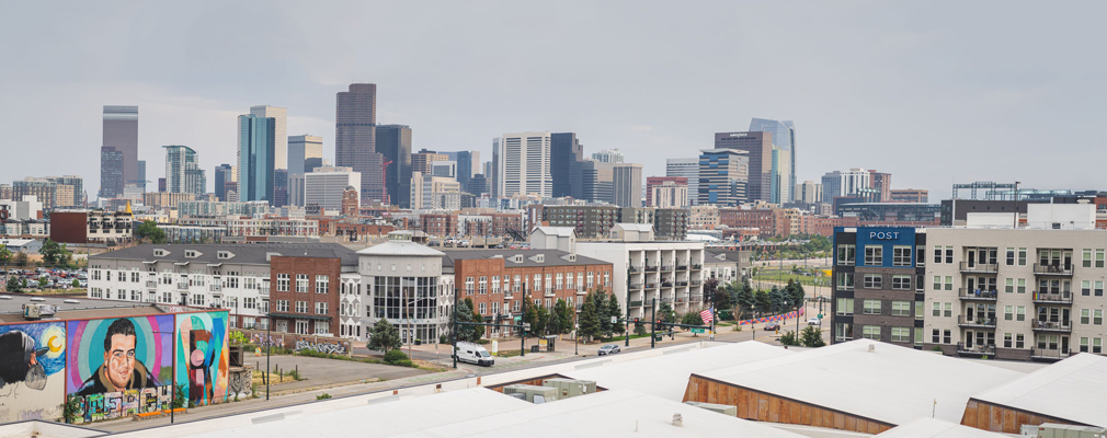 Panoramic photograph of several multistory buildings, one of which has a wall of murals, at an intersection in the RiNo Art District, with the downtown skyline in the background. 