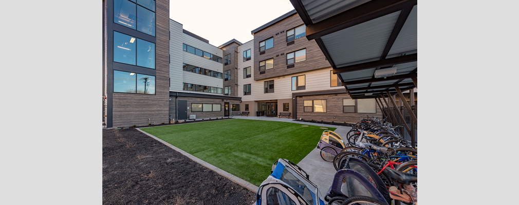 Photograph of a grassed courtyard framed by a four-story multifamily building, with several bicycles on the right edge of the photograph parked under a canopy.