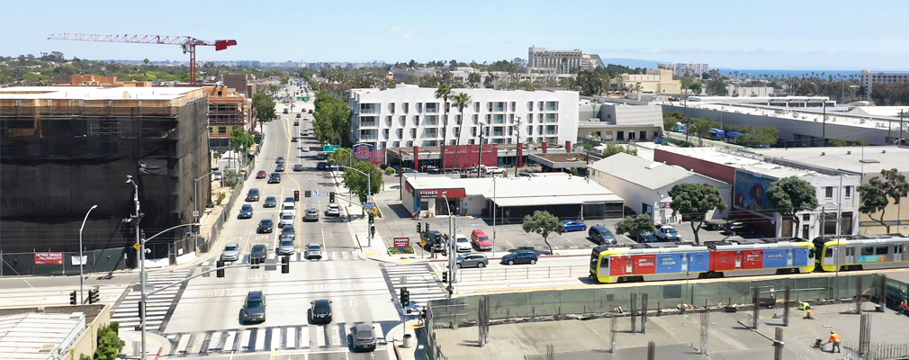 Photograph of a multistory residential building in the middle of the picture, with numerous other buildings nearby and two intersecting thoroughfares, one of which includes a light rail line.