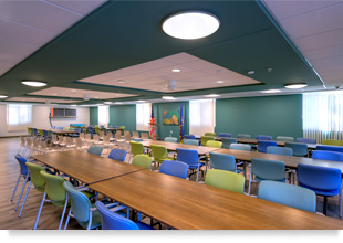Room with three rows of chairs and desks with two flags in the background, windows, and curtains.