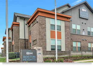 The street façade of a two-story apartment building, with a wall sign in the gable of the building and a monument sign in the foreground reading “BH Boston Heights.”