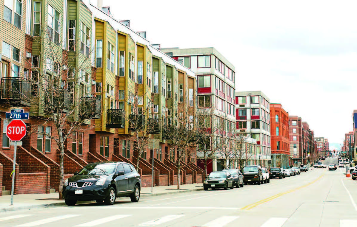 View of three- and four-story buildings fronting a two-way street with parked cars.