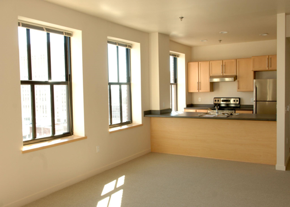 Interior of an apartment showing the kitchen with three large windows.