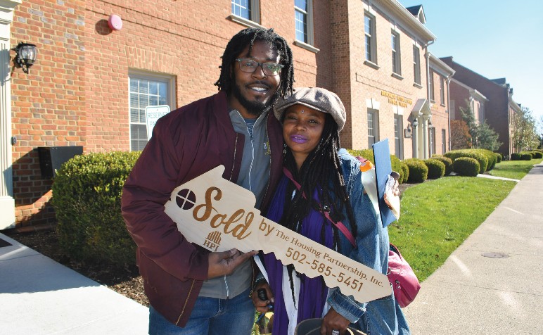A man and a woman posing in front of a two-story brick building and holding a large wooden key with the words "Sold by The Housing Partnership Inc." and a phone number on it.
