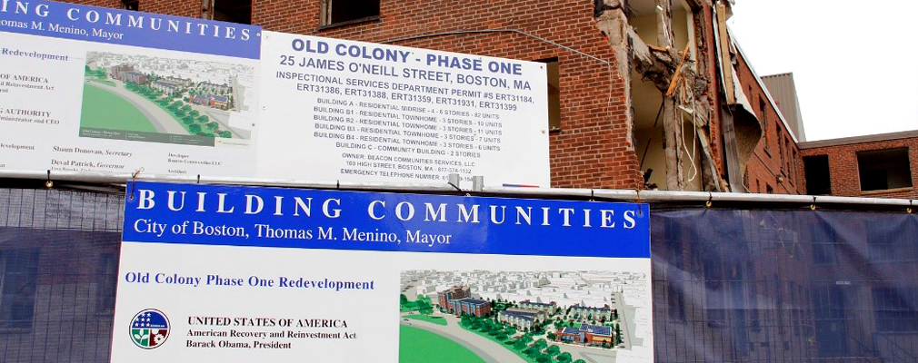 Photograph of a partially demolished multistory brick building with two large signs in the foreground detailing the redevelopment plans for Old Colony Homes Phase I.