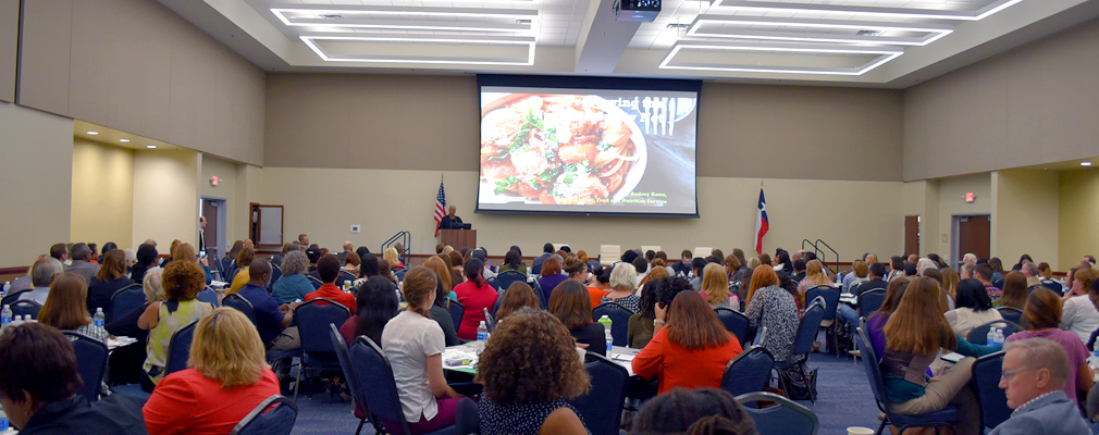 Photograph of dozens of attendees at the 2016 Dallas Hunger Summit seated at tables in a large meeting room and watching a presentation.