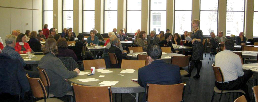 Photograph of people sitting at a dozen round tables and listening to a speaker in a large meeting room.