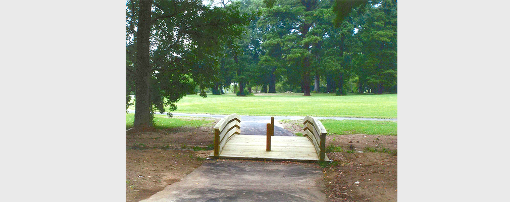 Photograph of a wooden bicycle/pedestrian bridge along a paved trail through an area of lawn and trees.