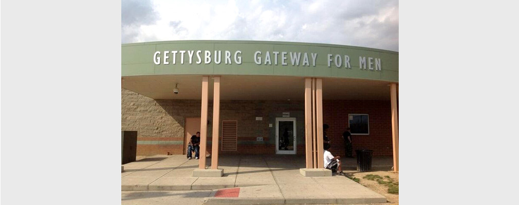 Photograph of the covered entrance to a one-story building with a sign reading “Gettysburg Gateway for Men” on the frieze at the roofline.
