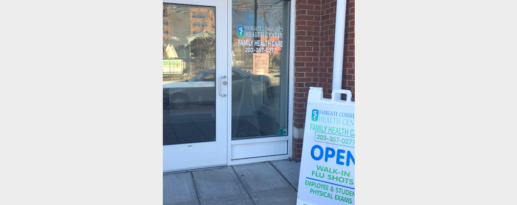 Photograph of the glazed entrance to a brick office with a window sign reading “Fairgate Community Health Center, Family Health Care” and with a sandwich board sign on the sidewalk advertising available services.   