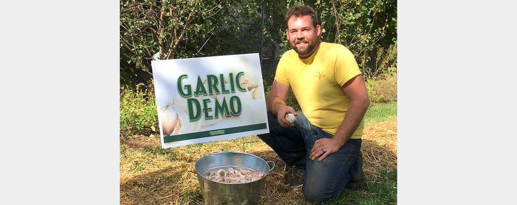 Photograph of a man kneeling next to a pail full of garlic bulbs, in front of a sign reading “Garlic Demo.”