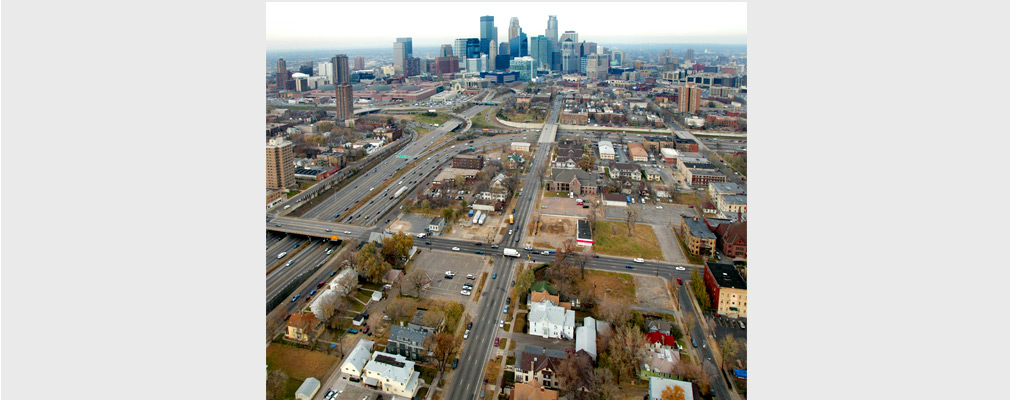 Low-altitude aerial photograph centered on an intersection of multilane streets, with downtown Minneapolis in the middleground. 
