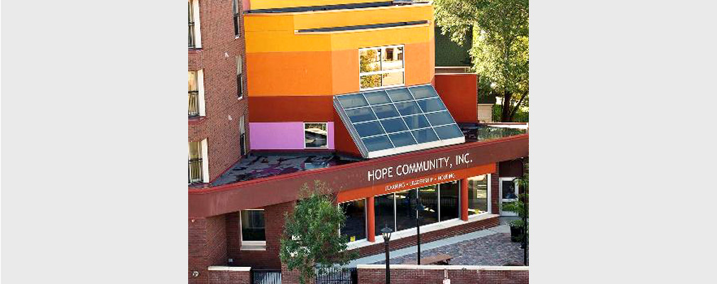 Photograph taken from several stories above ground of the corner entrance to a four-story residential building, with “Hope Community, Inc.” and “Learning · Leadership · Housing” above the entrance. 