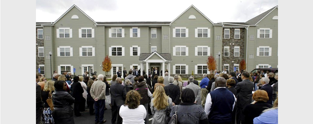 Photograph of a large group of people at a ribbon-cutting ceremony for a three-story apartment building in the background.