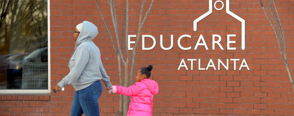 Photograph of a child holding her mother’s hand as they walk in front of a building with the sign “Educare Atlanta.”