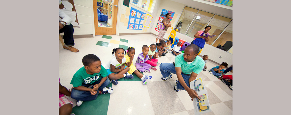 Photograph of five children sitting on the floor of a classroom as an instructor points to an opened book, with other adults and children in the background. 